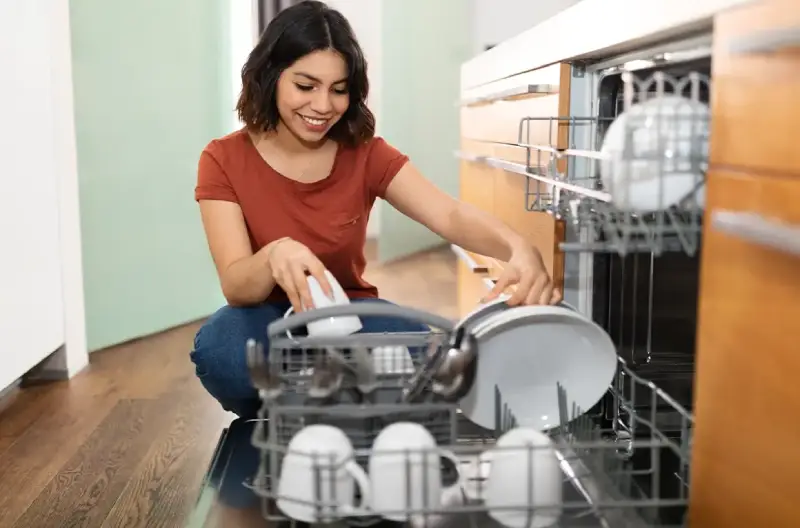 A woman wearing a red shirt and jeans is smiling while placing a mug and plate into a dishwasher.